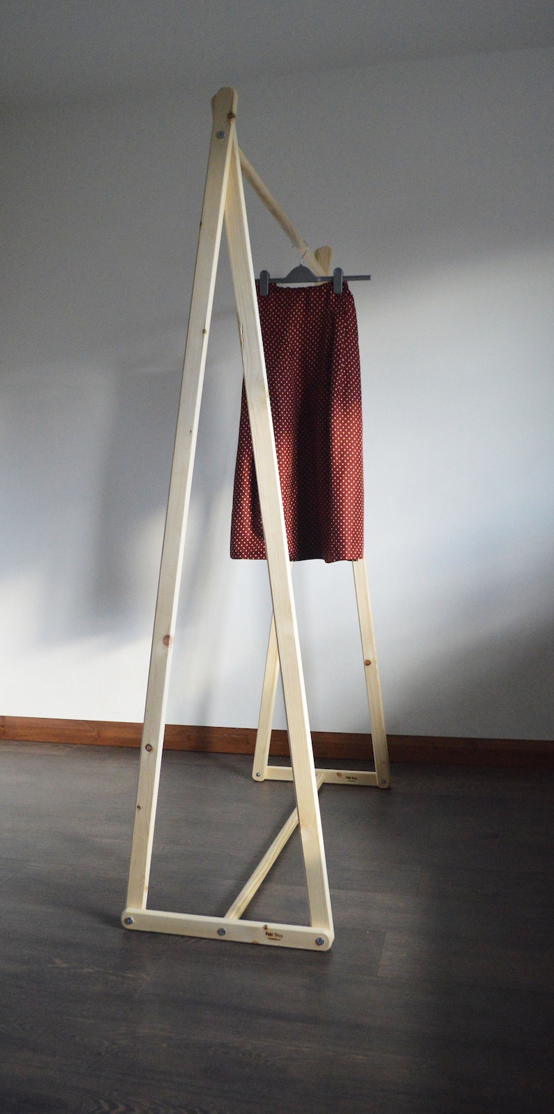May include: A wooden clothing rack with a skirt hanging on a hanger. The skirt is a dark red color with small white polka dots. The rack is made of light-colored wood and has an A-frame design.