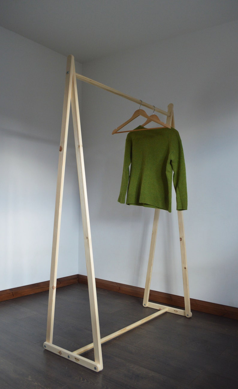May include: A wooden clothing rack with a simple A-frame design. A green sweater hangs on a wooden hanger. The rack is made of light-colored wood and stands on a dark wood floor.