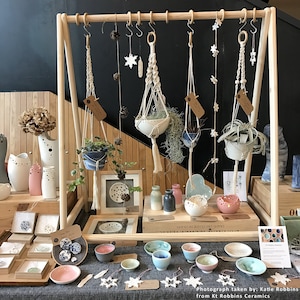 Könnte beinhalten: Eine Ausstellung handgefertigter Keramik und Makramee-Pflanzenaufhänger. Die Ausstellung umfasst kleine Schalen, Vasen und Hängepflanzgefäße mit Grün. Dekorative Elemente wie Schneeflocken und Tannenzapfen sind ebenfalls zu sehen. Die Farbpalette umfasst Weiß, Blau, Rosa und Naturtöne.