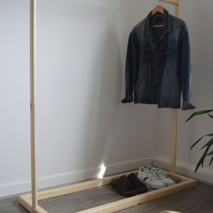 Könnte beinhalten: Kleiderständer aus hellem Holz mit einer Jeansjacke an einem Holzbügel. Braune Sandalen und weiße Turnschuhe stehen auf der Basis. Der Ständer steht vor einer weißen Wand.