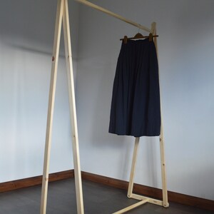 May include: A wooden clothing rack with a navy blue skirt hanging on a wooden hanger. The rack is made of light-colored wood and has a simple, minimalist design. The skirt is pleated and appears to be made of a soft fabric.