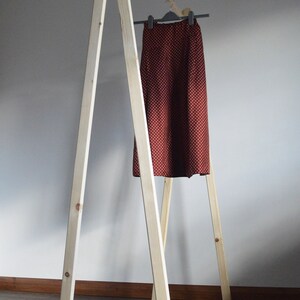 May include: A-frame wooden clothing rack with a skirt hanging on a hanger. The skirt is a dark red color with a white polka dot pattern. The rack is made of light-colored wood and is assembled with screws.