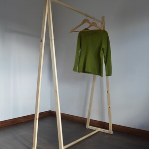 May include: A wooden clothing rack with a simple A-frame design. A green sweater hangs on a wooden hanger. The rack is made of light-colored wood and stands on a dark wood floor.