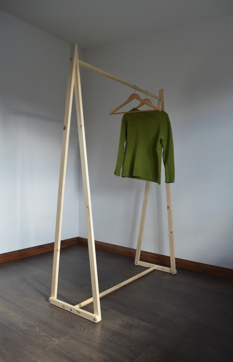 May include: A wooden clothing rack with a simple A-frame design. A green sweater hangs on a wooden hanger. The rack is made of light-colored wood and stands on a dark wood floor.
