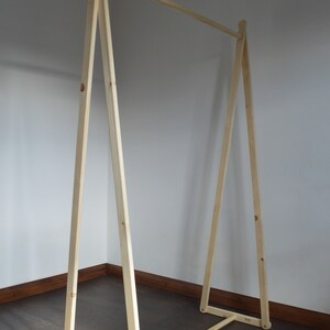 May include: A wooden clothing rack with a simple A-frame design. The natural wood rack has a horizontal bar for hanging clothes and is approximately 60 inches wide and 60 inches tall. The rack is constructed with visible screws and joints.