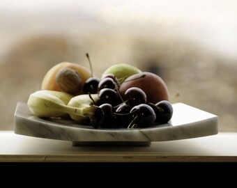 Op de afbeelding: Een witte marmeren kom gevuld met fruit, waaronder bananen, kersen en een perzik. De kom staat op een houten oppervlak.