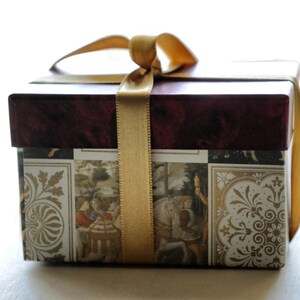 Op de afbeelding: Een decoratieve geschenkdoos met een bordeauxrode deksel en een patroonbasis met historische sc&egrave;nes en gouden accenten. De doos is vastgebonden met een gouden lint, wat een klassieke en elegante presentatie cre&euml;ert.