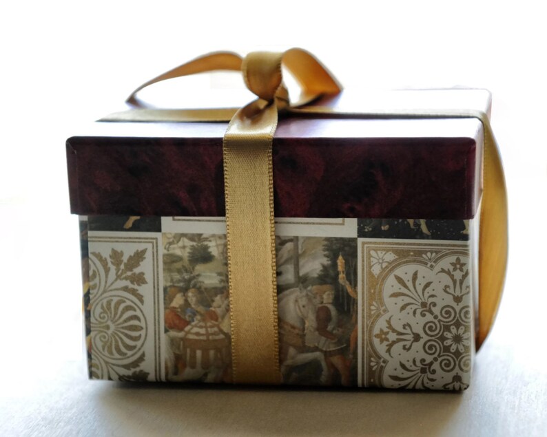 Op de afbeelding: Een decoratieve geschenkdoos met een bordeauxrode deksel en een patroonbasis met historische sc&egrave;nes en gouden accenten. De doos is vastgebonden met een gouden lint, wat een klassieke en elegante presentatie cre&euml;ert.