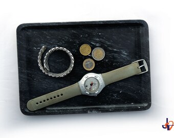 Op de afbeelding: Een zwarte marmeren tray met een zilveren horloge met een grijze band, een zilveren armband en verschillende euromunten. Het horloge heeft een zilveren wijzerplaat en een grijze band. De armband heeft een opgerold ontwerp.