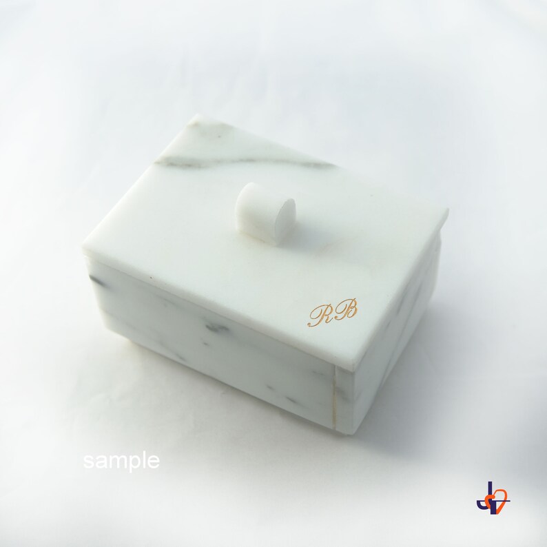 Op de afbeelding: Een witte marmeren doos met een deksel en een kleine, afgeronde knop. De doos heeft grijze adering en goudkleurige initialen "RB" aan de zijkant. Het woord "sample" staat linksonder afgedrukt.