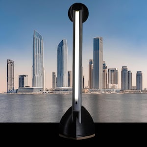 Può includere: Una moderna lampada da terra nera e argento con una striscia di luce bianca verticale. La lampada ha una parte superiore circolare e una base triangolare. Lo sfondo presenta un paesaggio urbano con edifici alti e uno specchio d'acqua.