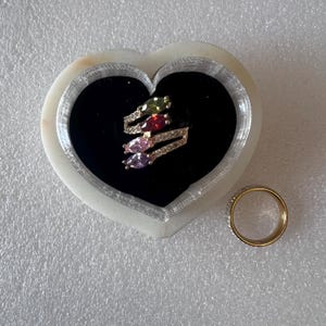 May include: A gold ring with a band of small clear stones is next to a heart-shaped jewellery box. The box contains a ring with four colourful gemstones: green, red, pink, and purple. The jewellery box is white with a black interior.