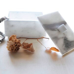 Op de afbeelding: Een witte marmeren doos met een deksel, met grijze adering en een kleine witte vierkante knop. Een zilverkleurige armband en gedroogde bloemelementen zijn in de buurt gerangschikt, wat een minimalistische esthetiek cre&euml;ert.