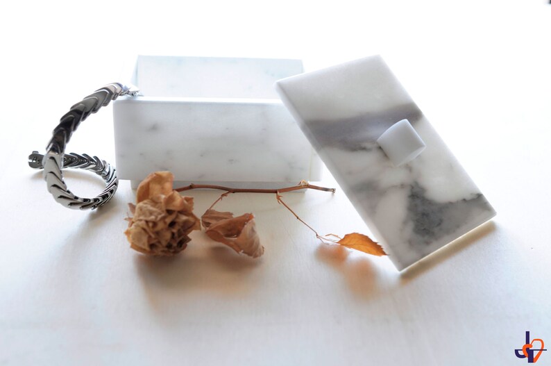 Op de afbeelding: Een witte marmeren doos met een deksel, met grijze adering en een kleine witte vierkante knop. Een zilverkleurige armband en gedroogde bloemelementen zijn in de buurt gerangschikt, wat een minimalistische esthetiek cre&euml;ert.