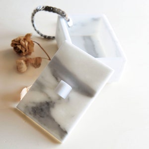 Op de afbeelding: Een witte marmeren doos met een scharnierend deksel, met grijze adering. Het deksel staat iets open en onthult de binnenkant. Een zilveren armband en een gedroogde bloem liggen naast de doos. De doos is een decoratief opbergitem.