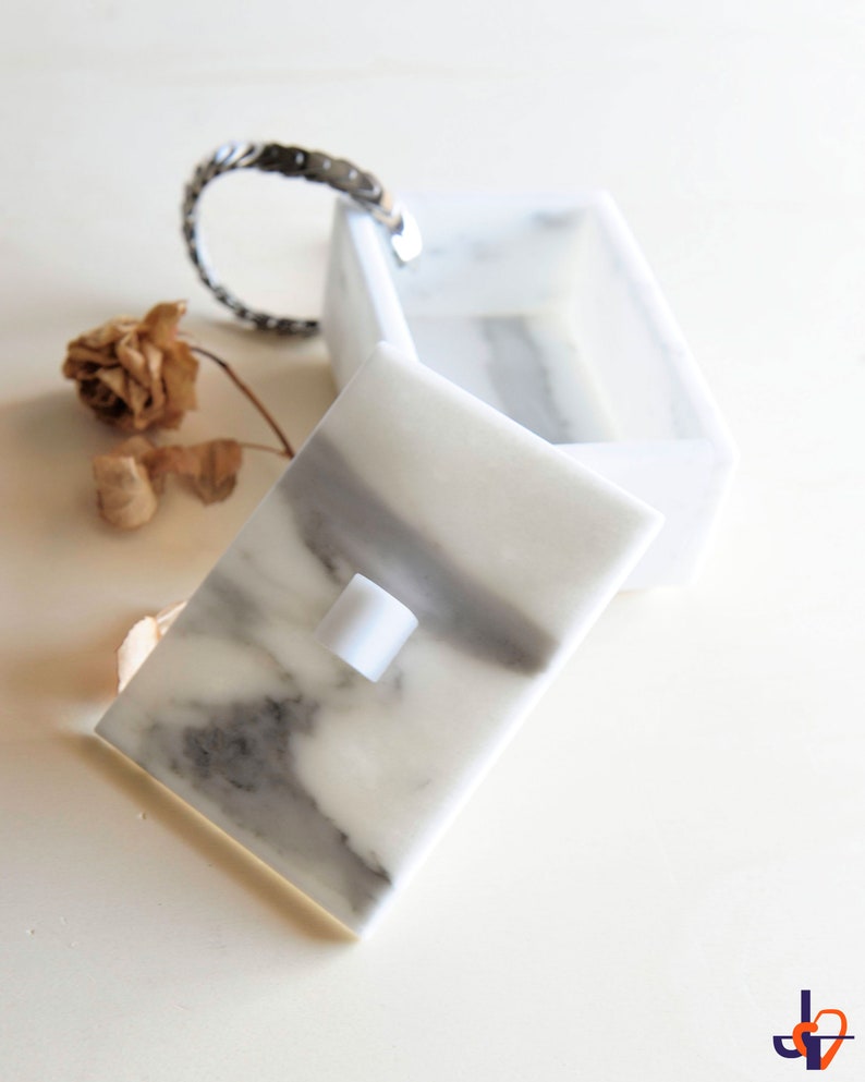 Op de afbeelding: Een witte marmeren doos met een scharnierend deksel, met grijze adering. Het deksel staat iets open en onthult de binnenkant. Een zilveren armband en een gedroogde bloem liggen naast de doos. De doos is een decoratief opbergitem.
