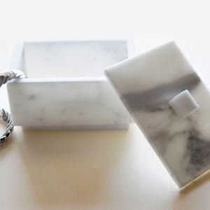 Op de afbeelding: Een rechthoekige witte marmeren doos met een deksel, met grijze adering. Het deksel heeft een kleine, vierkante handgreep. Een zilverkleurige, gevlochten ring is aan de doos bevestigd. De doos is open en toont de binnenkant.