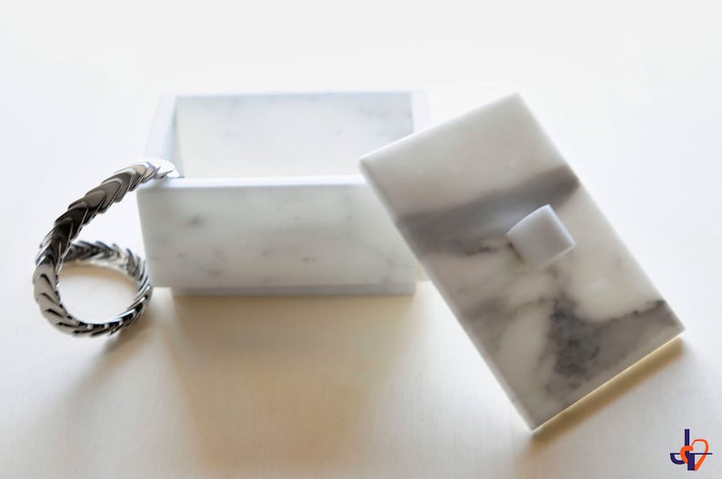 Op de afbeelding: Een rechthoekige witte marmeren doos met een deksel, met grijze adering. Het deksel heeft een kleine, vierkante handgreep. Een zilverkleurige, gevlochten ring is aan de doos bevestigd. De doos is open en toont de binnenkant.