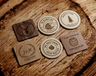 Könnte beinhalten: Eine Sammlung von sechs Holzuntersetzern mit eingravierten Designs. Die Untersetzer zeigen verschiedene Logos und Texte, darunter "The Iron Stag", "Oceanside Brewing Co.", "North Pine Brewing Co.", "The Daily Grind Coffee Roasters" und "Stone Creek Cellars". Ein Untersetzer zeigt "J&M" und ein Datum.