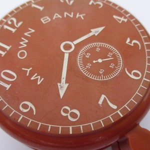 Puede incluir: Un reloj de plástico rojo con números y manecillas blancas. La esfera del reloj dice "My Own Bank".