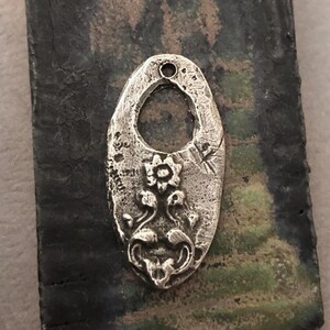 May include: A silver-colored, oval-shaped pendant with a textured surface and a floral design. The pendant has a central opening and a small hole at the top for a chain. The floral design includes a central flower and stylized leaves.