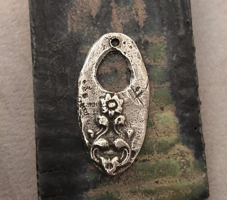 May include: A silver-colored, oval-shaped pendant with a textured surface and a floral design. The pendant has a central opening and a small hole at the top for a chain. The floral design includes a central flower and stylized leaves.