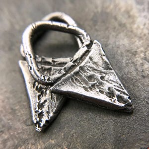 May include: A silver metal pendant with a textured surface and a loop at the top. The pendant is shaped like a triangle with a rounded point.
