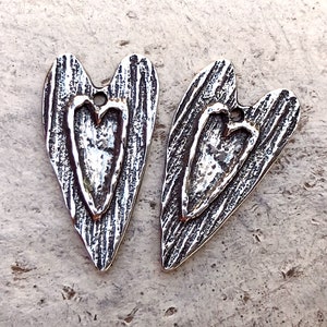 Op de afbeelding: Twee zilveren hartvormige bedels met een getextureerd oppervlak en een kleiner hartontwerp in elk. De bedels hebben een gat aan de bovenkant om aan sieraden te bevestigen.
