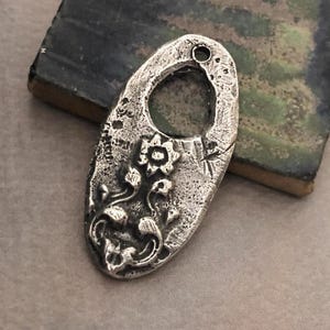 May include: Oval-shaped, silver-toned pendant with a floral design. The pendant features a textured surface and a large central opening. The floral design includes a flower and leaves. A small hole is at the top for stringing.