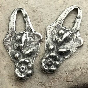 Op de afbeelding: Twee zilverkleurige metalen bedels met een bloemmotief. Elke bedel heeft een lus aan de bovenkant en een gedetailleerd bloemmotief met een centrale bloem en bladeren.