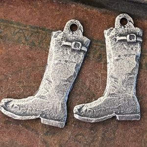 May include: Two silver-toned metal charms shaped like boots. The boots have a textured surface and a buckle detail.