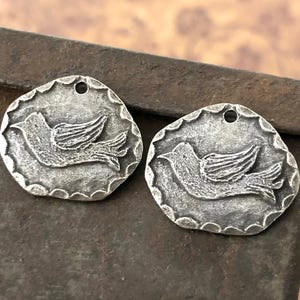 Op de afbeelding: Twee zilverkleurige metalen bedels in de vorm van een vogel met een gat aan de bovenkant om op te hangen. De bedels hebben een getextureerd oppervlak en een licht verweerde uitstraling.