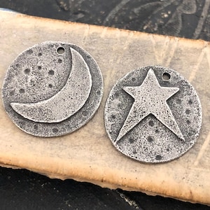 May include: Two circular silver-toned metal charms with a weathered, textured finish. One charm features a raised crescent moon design, while the other displays a raised five-pointed star. Both pieces have a small hole at the top for hanging.