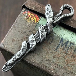 Könnte beinhalten: Ein silberfarbener Anhänger in Form einer Schlange, die sich um einen Stab windet. Die Schlange hat eine detaillierte Textur, und der Anhänger hat eine Schlaufe oben. Der Hintergrund ist eine verwitterte Metallbox.