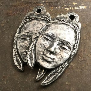 May include: Two silver-toned metal pendants featuring detailed face sculptures with closed eyes. Each pendant has a braided design around the head and feathers on the sides.