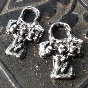 Puede incluir: Dos abalorios de metal plateado en forma de cruz con un bucle en la parte superior. La cruz tiene una superficie texturizada y peque&ntilde;os puntos en relieve.