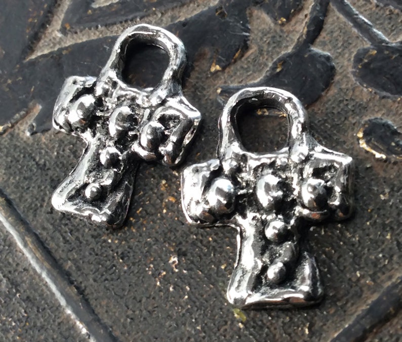 Puede incluir: Dos abalorios de metal plateado en forma de cruz con un bucle en la parte superior. La cruz tiene una superficie texturizada y peque&ntilde;os puntos en relieve.