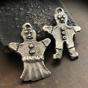 May include: Two silver-toned gingerbread man charms. Each charm has a loop at the top for hanging, a smiling face, and button details. The charms are detailed with textured lines and are set against a dark, textured background.