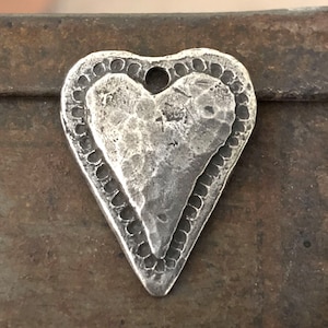May include: A silver heart-shaped pendant with a textured surface and a small hole at the top. The pendant has a dotted border and a hammered appearance, suggesting a handcrafted design.