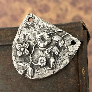 Puede incluir: Un colgante de metal plateado con un dise&ntilde;o floral. El colgante presenta una superficie texturizada y dos agujeros para colgar. El dise&ntilde;o incluye una flor, hojas y bayas.