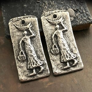 May include: Two silver-toned metal charms featuring a raised design of a stylised figure carrying a basket. The charms have a textured surface and a rectangular shape.