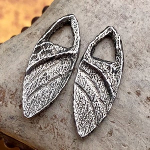 May include: Two silver metal pendants shaped like leaves with a textured surface. Each pendant has a hole in the center.