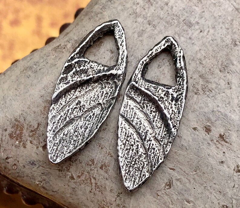 May include: Two silver metal pendants shaped like leaves with a textured surface. Each pendant has a hole in the center.