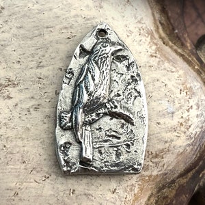 May include: A silver pendant shaped like a bird perched on a branch. The pendant has a textured surface and a detailed design.