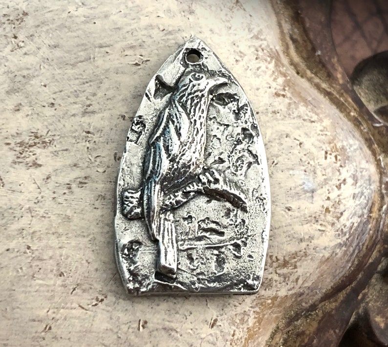 May include: A silver pendant shaped like a bird perched on a branch. The pendant has a textured surface and a detailed design.