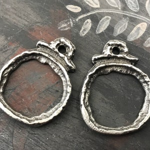 May include: Two silver-toned metal charms in the shape of a circle with a raised top. The charms have a rough, textured surface and a hole in the top for attaching to a necklace or bracelet.