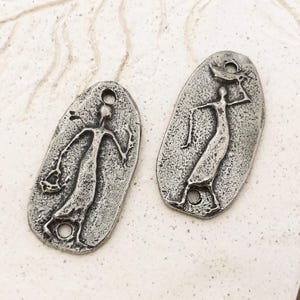 Peut inclure: Deux pendentifs en m&eacute;tal argent&eacute; de forme ovale. Chaque pendentif repr&eacute;sente une figure stylis&eacute;e aux bras tendus. Les pendentifs ont une surface textur&eacute;e et un trou en haut et en bas pour l'enfilage.
