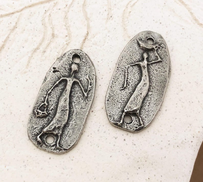 Peut inclure: Deux pendentifs en m&eacute;tal argent&eacute; de forme ovale. Chaque pendentif repr&eacute;sente une figure stylis&eacute;e aux bras tendus. Les pendentifs ont une surface textur&eacute;e et un trou en haut et en bas pour l'enfilage.