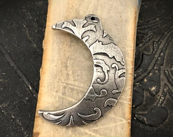 May include: A crescent moon pendant made of antiqued silver-toned metal, featuring an intricate embossed scrollwork pattern across its surface and a small circular hole at the top for attachment to a chain or cord.
