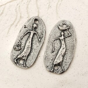 Peut inclure: Deux pendentifs argent&eacute;s de forme ovale, perc&eacute;s d'un trou en haut et en bas. Chaque pendentif repr&eacute;sente une figure stylis&eacute;e aux bras tendus. L'une tient un objet, l'autre porte une forme de croissant sur la t&ecirc;te.